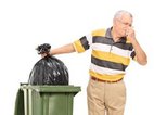 Een oudere man knijpt zijn neus dicht als hij een vuilniszak in de afvalcontainer laat zakken zonder uitstelgedrag.
