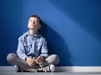 De nadenkende autistische jongen zit met gekruiste benen op witte vloer tegen blauwe muur