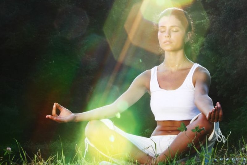 Een vrouw zit heel stil te mediteren in het gras. De achtergrond is donker, daardoor zie je dat ze in de schitterende lichtstralen van de zon zit