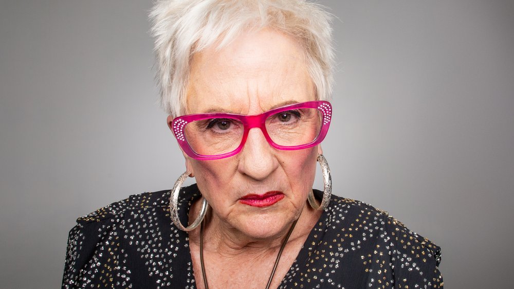 Een oudere narcistische vrouw met een rode bril op kijkt afkeurend, dwingend
