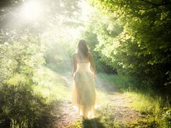 Een hooggevoelige en hoogsensitieve vrouw in een fladderende witte jurk loopt in de zonnestralen, over een pad door een lichtgroen bos