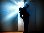 In een donkere kamer gluurt een man achter een deur die op een kier staat. In de ruimte achter de deur is het licht.