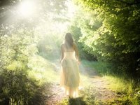 Een vrouw in een fladderende witte jurk loopt in de zonnestralen, over een pad door een lichtgroen bos
