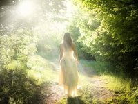 Een vrouw in een fladderende witte jurk loopt in de zonnestralen, over een pad door een lichtgroen bos