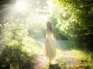 Een hooggevoelige en hoogsensitieve vrouw in een fladderende witte jurk loopt in de zonnestralen, over een pad door een lichtgroen bos