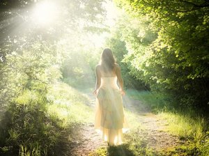 Een hooggevoelige en hoogsensitieve vrouw in een fladderende witte jurk loopt in de zonnestralen, over een pad door een lichtgroen bos