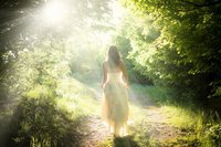 Een hooggevoelige en hoogsensitieve vrouw in een fladderende witte jurk loopt in de zonnestralen, over een pad door een lichtgroen bos