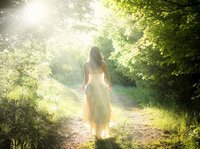 Een hooggevoelige en hoogsensitieve vrouw in een fladderende witte jurk loopt in de zonnestralen, over een pad door een lichtgroen bos