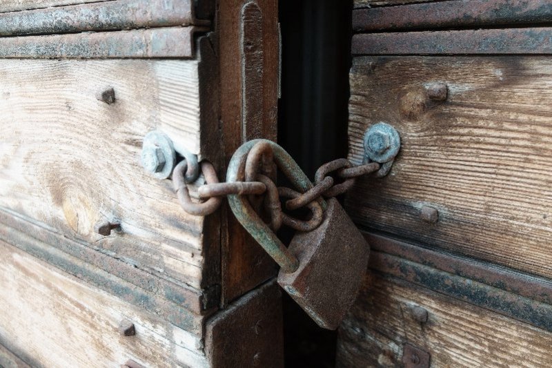 Een ketting met een hangstot op een oude deur van een schuur. De deur staat op een kier en binnen is het helemaal donker.