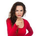 Een vrouw met een rood shirt aan kijkt boos naar de camera. Een hand in de zij, en met de andere wijst ze naar de camera.