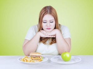 Een vrouw zit aan tafel. Ze moet kiezen tussen een bord gezonde voeding en een bord met hamburger en patat.