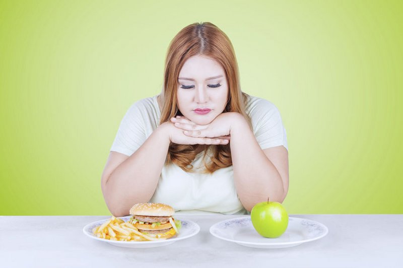Een vrouw zit aan tafel. Ze wil afvallen en moet kiezen tussen een bord gezonde voeding en een bord met hamburger en patat.