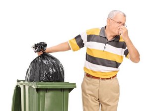 Een oudere man knijpt zijn neus dicht terwijl hij een vuilniszak in de container laat zakken.