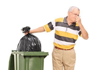 Een oudere man knijpt zijn neus dicht terwijl hij een vuilniszak in de container laat zakken.