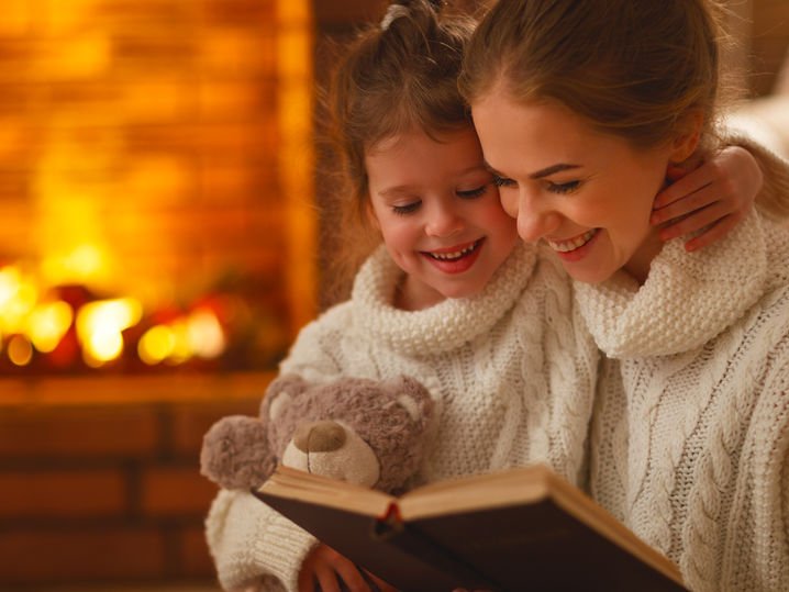 Een moeder heeft een kind op schoot en leest een boek voor, warm en knus bij de open haard.