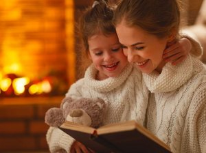 Een moeder heeft een kind op schoot en leest een boek voor, warm en knus bij de open haard.