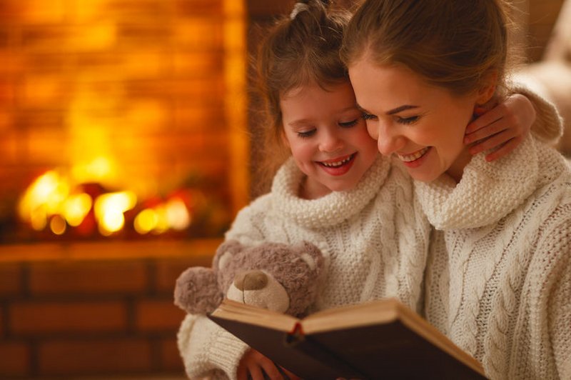 Een moeder heeft een kind op schoot en leest een boek voor, warm en knus bij de open haard.