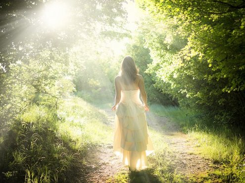 Een hooggevoelige en hoogsensitieve vrouw in een fladderende witte jurk loopt in de zonnestralen, over een pad door een lichtgroen bos