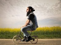 Een hipster met baard en knotje fietst op een veel te klein fietsje langs een veld