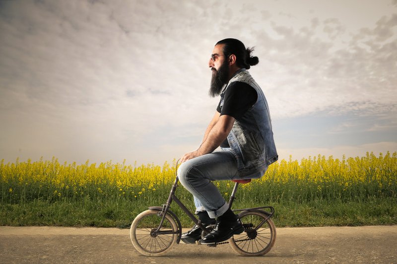 Een hipster met baard en knotje fietst op een veel te klein fietsje langs een veld
