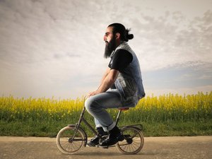 Een hipster met baard en knotje fietst op een veel te klein fietsje langs een veld